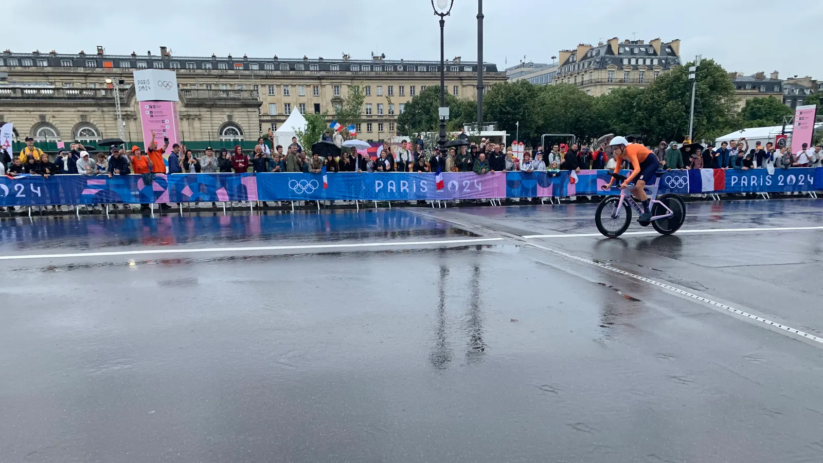 Coureuse en maillot orange seule sur une chaussée mouillée, devant des spectateurs derrière des barrières