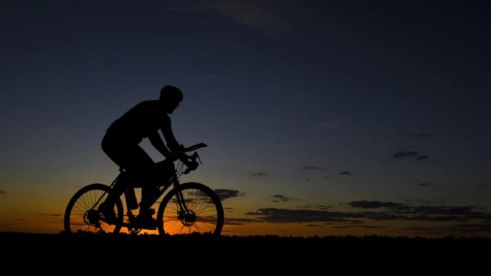Silhouette d'un cycliste sur sa route au coucher du soleil, ciel bleu nuit et horizon orange