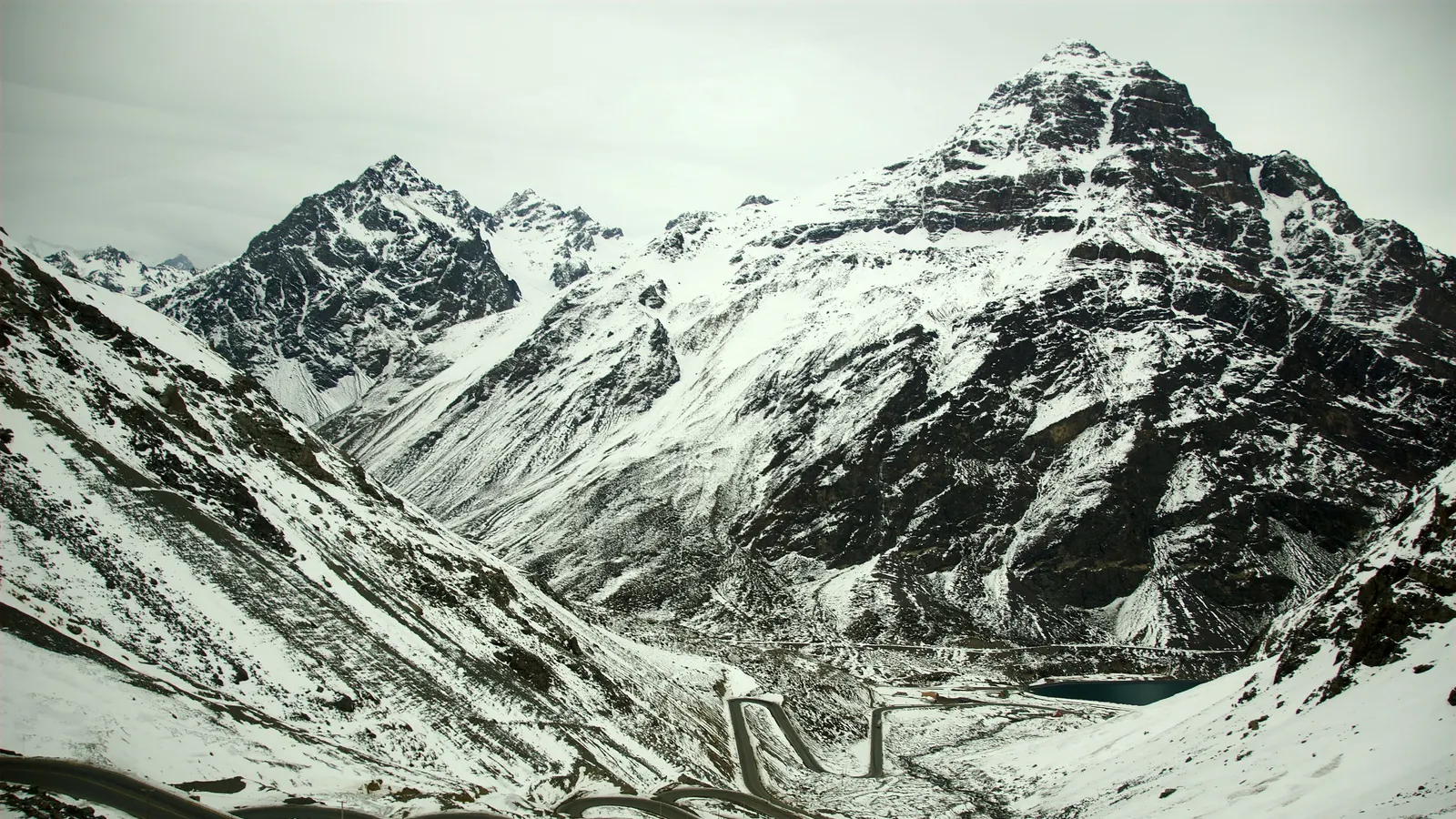 Route en lacets au fond d'une vallée de haute montagne enneigée, sous un ciel gris