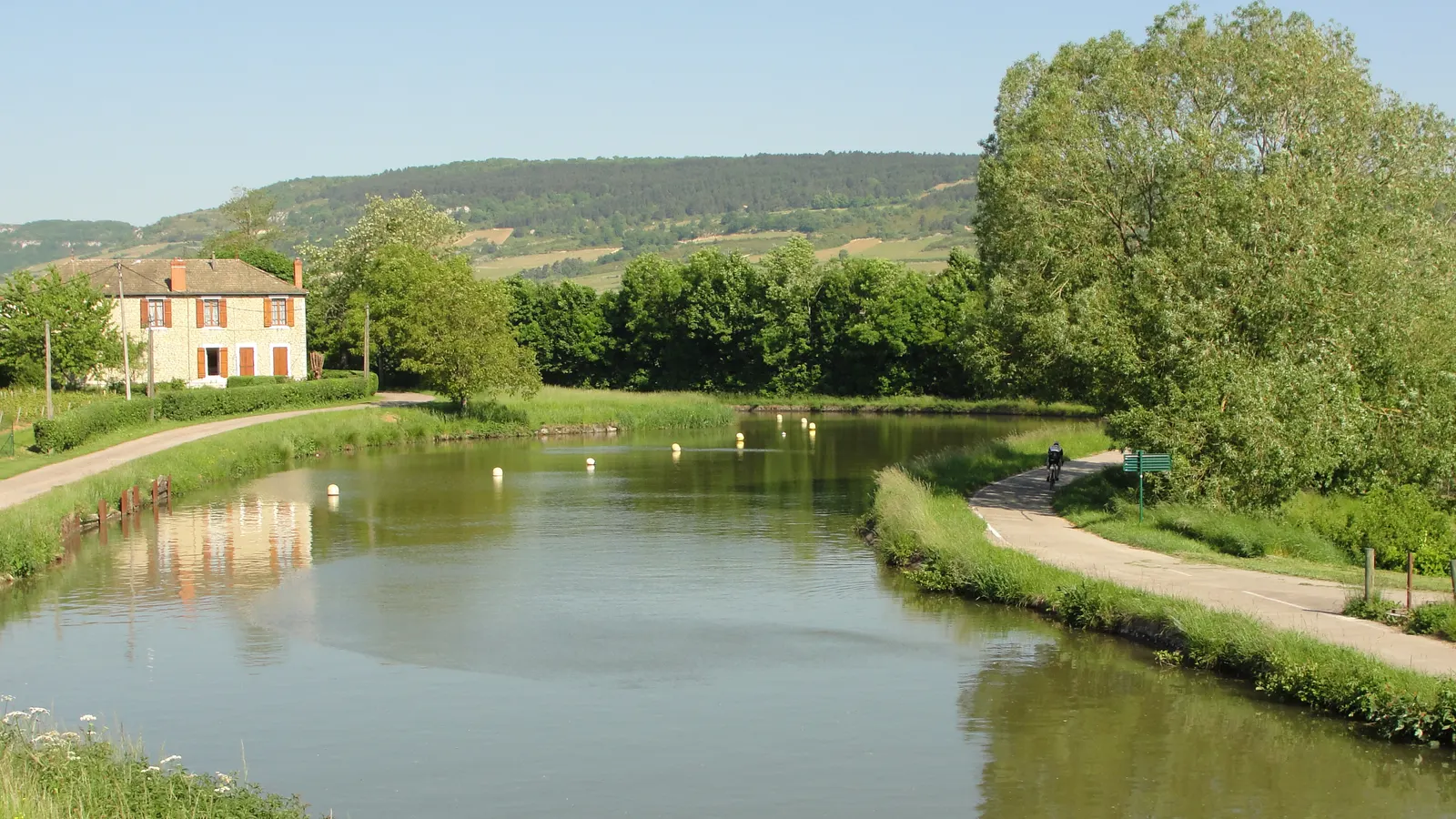 Canal bordé d'une piste cyclable, avec une maison de pierre et des coteaux de vignes à l'arrière-plan